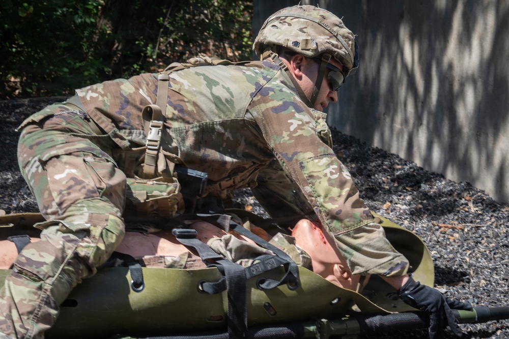A U.S. Army soldier in Operational Camouflage Pattern (OCP) uniform and combat helmet is performing a casualty evacuation drill, loading or securing a simulated casualty onto a litter/stretcher. The soldier appears to be conducting tactical combat casualty care (TCCC) training, as evidenced by the training mannequin or simulated casualty with visible simulated wounds. The setting appears to be an outdoor training environment.