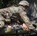 A U.S. Army soldier in Operational Camouflage Pattern (OCP) uniform and combat helmet is performing a casualty evacuation drill, loading or securing a simulated casualty onto a litter/stretcher. The soldier appears to be conducting tactical combat casualty care (TCCC) training, as evidenced by the training mannequin or simulated casualty with visible simulated wounds. The setting appears to be an outdoor training environment.