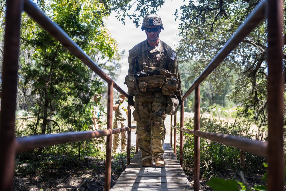 A soldier in full combat gear and camouflage uniform crosses a narrow wooden footbridge with rusted metal railings in a wooded outdoor environment. The soldier is carrying a heavy tactical load including a rucksack and equipment pouches, wearing a helmet and sunglasses. At least one additional soldier is visible in the background following the same path.
