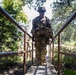 A soldier in full combat gear and camouflage uniform crosses a narrow wooden footbridge with rusted metal railings in a wooded outdoor environment. The soldier is carrying a heavy tactical load including a rucksack and equipment pouches, wearing a helmet and sunglasses. At least one additional soldier is visible in the background following the same path.
