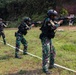 A group of military or law enforcement personnel in camouflage uniforms and black tactical gear are conducting a live-fire shooting exercise on an outdoor range. Multiple shooters are aligned in a row, aiming rifles at paper human-silhouette targets mounted on wooden stands. Smoke is visible near the targets, indicating active firing.