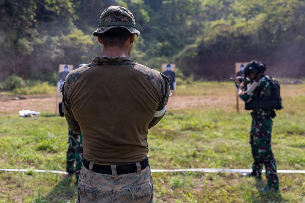 The image depicts a military shooting range exercise with at least two personnel visible. One soldier in US Marine Corps MARPAT digital camouflage uniform observes from behind a safety line, while another soldier in a different camouflage pattern (green/brown tropical) actively engages paper silhouette targets downrange. The setting appears to be an outdoor range with natural vegetation in the background, suggesting a joint training exercise.