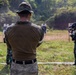 The image depicts a military shooting range exercise with at least two personnel visible. One soldier in US Marine Corps MARPAT digital camouflage uniform observes from behind a safety line, while another soldier in a different camouflage pattern (green/brown tropical) actively engages paper silhouette targets downrange. The setting appears to be an outdoor range with natural vegetation in the background, suggesting a joint training exercise.