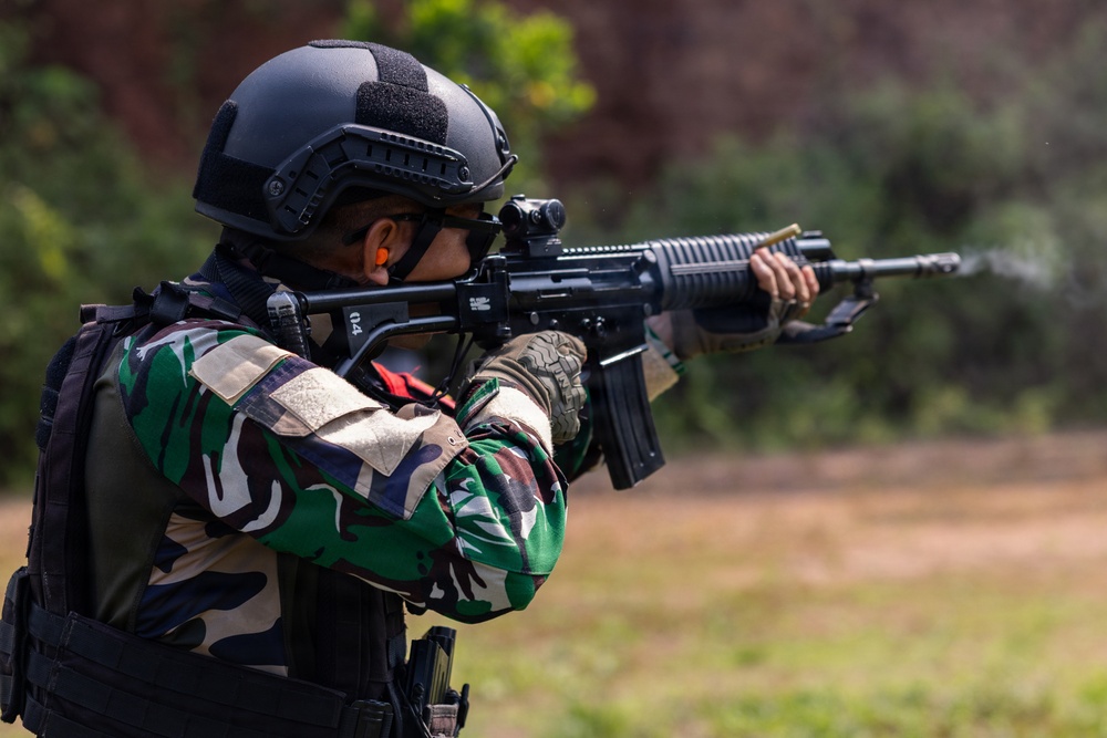 A soldier or military/law enforcement personnel is shown in a shooting stance, aiming a rifle downrange. The individual is wearing full tactical gear including a ballistic helmet, camouflage uniform, body armor vest, and tactical gloves. The setting appears to be an outdoor shooting range or training area.