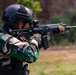 A soldier or military/law enforcement personnel is shown in a shooting stance, aiming a rifle downrange. The individual is wearing full tactical gear including a ballistic helmet, camouflage uniform, body armor vest, and tactical gloves. The setting appears to be an outdoor shooting range or training area.