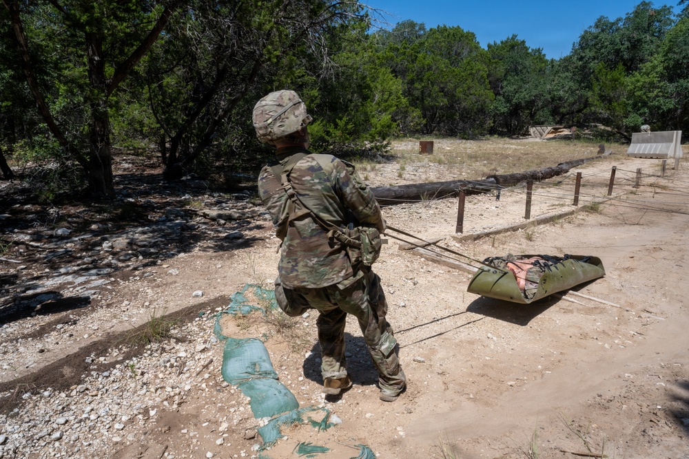 A soldier in full MultiCam/OCP camouflage uniform is dragging an improvised litter or casualty evacuation sled across a sandy, arid training area. The scene appears to be a military field training exercise focused on combat casualty care or MEDEVAC procedures. The environment suggests a southwestern U.S. military installation with scrub trees and rocky terrain.