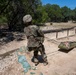 A soldier in full MultiCam/OCP camouflage uniform is dragging an improvised litter or casualty evacuation sled across a sandy, arid training area. The scene appears to be a military field training exercise focused on combat casualty care or MEDEVAC procedures. The environment suggests a southwestern U.S. military installation with scrub trees and rocky terrain.