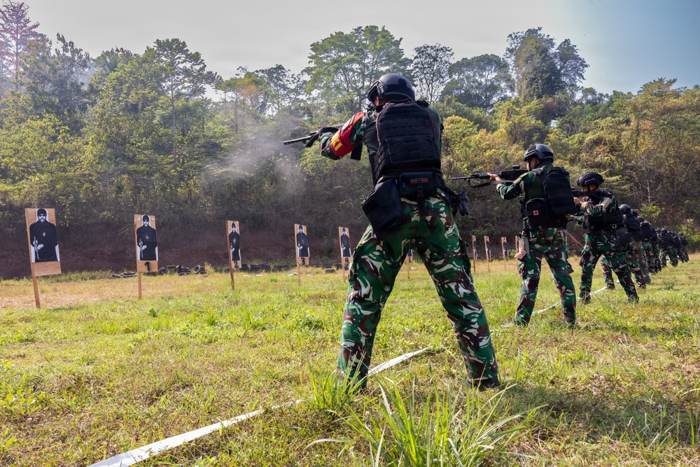 A group of military or law enforcement personnel are conducting a live-fire shooting exercise at an outdoor range. Multiple individuals in camouflage uniforms and black tactical gear are firing rifles at human-silhouette paper targets. Visible smoke from a discharged weapon is evident near the lead shooter in the foreground.