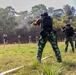 A group of military or law enforcement personnel are conducting a live-fire shooting exercise at an outdoor range. Multiple individuals in camouflage uniforms and black tactical gear are firing rifles at human-silhouette paper targets. Visible smoke from a discharged weapon is evident near the lead shooter in the foreground.