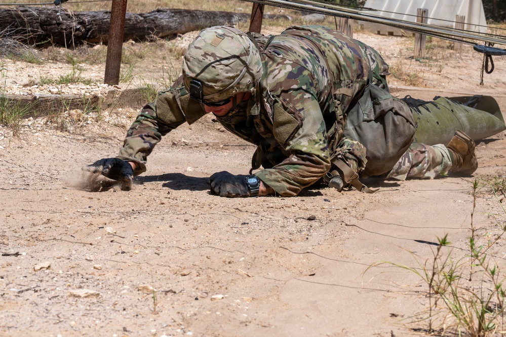 A soldier in full Operational Camouflage Pattern (OCP) uniform is low-crawling under an obstacle course wire barrier on a sandy, arid training ground. The soldier is wearing a combat helmet, tactical gloves, eye protection, and a watch, and appears to be actively pushing through the dirt with both hands. This appears to be a military training exercise, likely a confidence or obstacle course event.