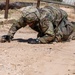 A soldier in full Operational Camouflage Pattern (OCP) uniform is low-crawling under an obstacle course wire barrier on a sandy, arid training ground. The soldier is wearing a combat helmet, tactical gloves, eye protection, and a watch, and appears to be actively pushing through the dirt with both hands. This appears to be a military training exercise, likely a confidence or obstacle course event.