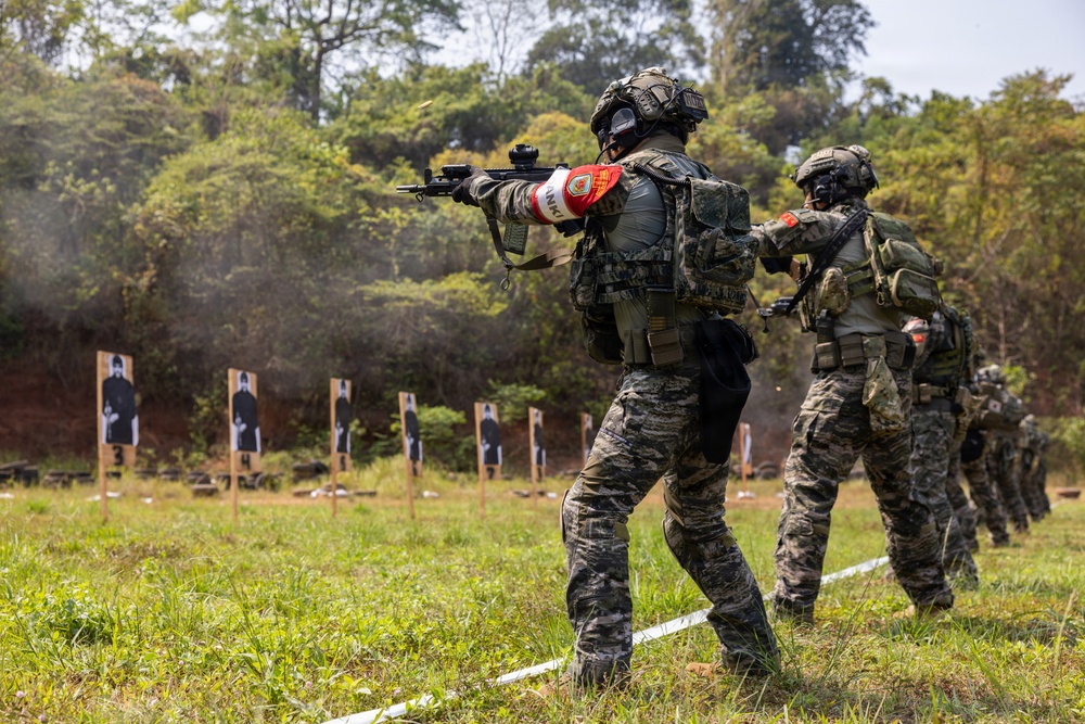 Military or law enforcement personnel are conducting a live-fire shooting range exercise in an outdoor, forested environment. Multiple armed individuals in camouflage uniforms and tactical gear are engaging silhouette targets downrange. The scene appears to be a structured training drill with at least one instructor guiding participants.