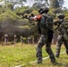 Military or law enforcement personnel are conducting a live-fire shooting range exercise in an outdoor, forested environment. Multiple armed individuals in camouflage uniforms and tactical gear are engaging silhouette targets downrange. The scene appears to be a structured training drill with at least one instructor guiding participants.