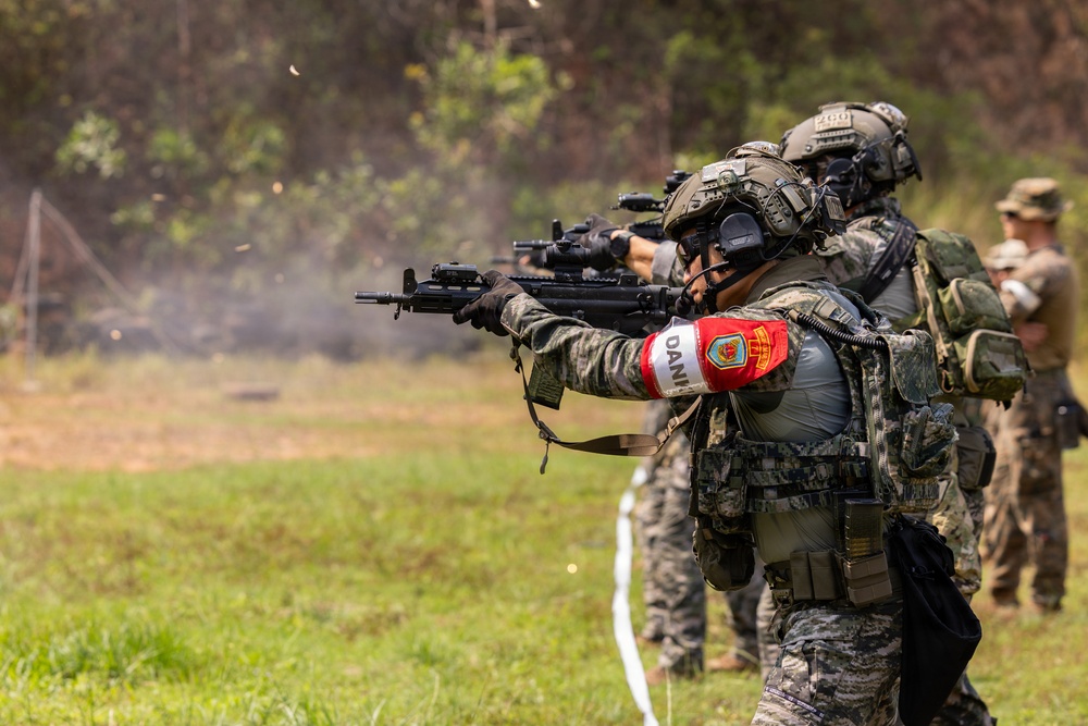 Military personnel are engaged in a live-fire training exercise on an outdoor range. The foreground soldier wears a red armband with visible insignia and text, suggesting a multinational or joint exercise. Smoke is visible near the muzzle of the lead weapon, indicating active firing.