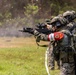 Military personnel are engaged in a live-fire training exercise on an outdoor range. The foreground soldier wears a red armband with visible insignia and text, suggesting a multinational or joint exercise. Smoke is visible near the muzzle of the lead weapon, indicating active firing.
