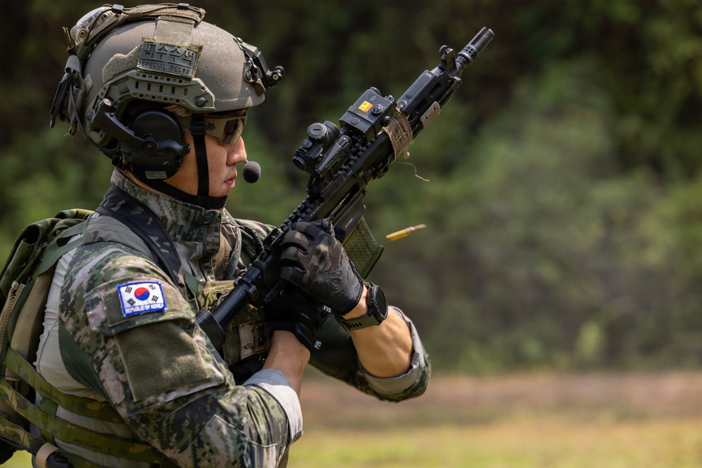 A Republic of Korea (ROK) Special Reconnaissance soldier is actively firing a rifle, with a spent brass casing visibly ejecting from the weapon. The soldier is equipped with full tactical gear including a ballistic helmet, communications headset, body armor, and optical sights. The setting appears to be an outdoor training or operational environment with a green, vegetated background.