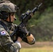 A Republic of Korea (ROK) Special Reconnaissance soldier is actively firing a rifle, with a spent brass casing visibly ejecting from the weapon. The soldier is equipped with full tactical gear including a ballistic helmet, communications headset, body armor, and optical sights. The setting appears to be an outdoor training or operational environment with a green, vegetated background.
