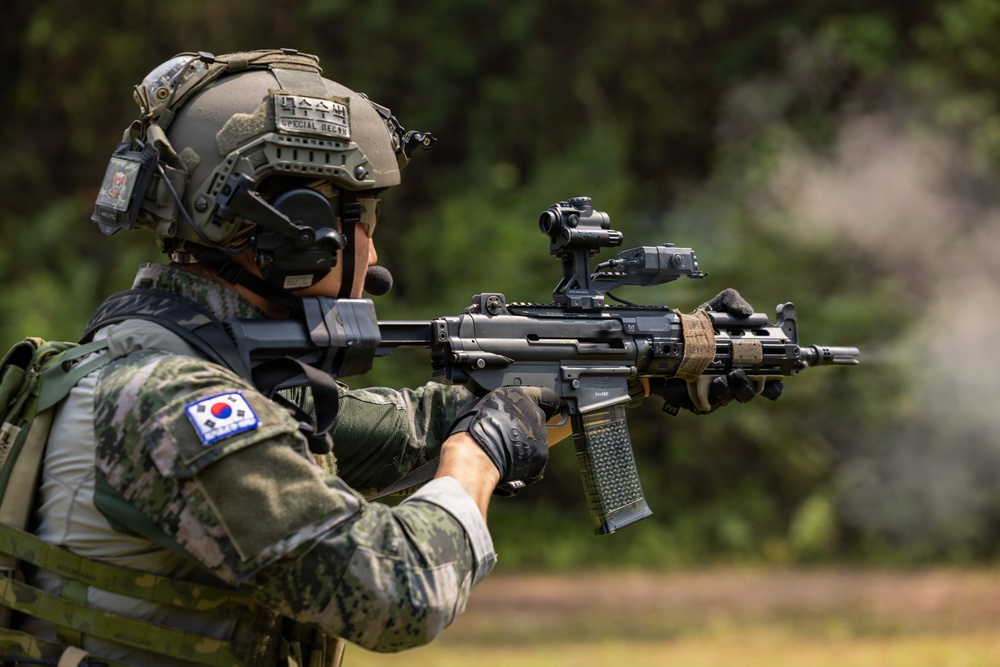 A South Korean special operations soldier is shown in a firing stance, aiming a rifle in an outdoor environment. The soldier is equipped with advanced tactical gear including a helmet with 'Special Recon' markings, communication headset, and a modern assault rifle with optics. The South Korean flag patch is clearly visible on the left shoulder of the camouflage uniform.