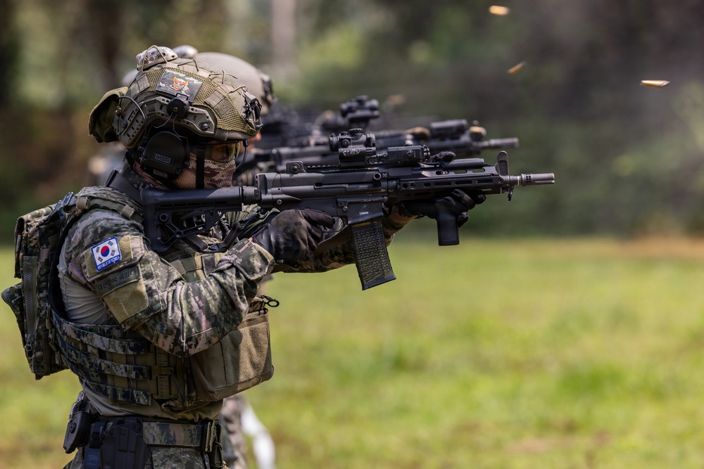 The image shows South Korean military personnel conducting a live-fire shooting exercise or demonstration. The foreground soldier is clearly identifiable by the South Korean flag patch on the left shoulder, firing what appears to be a Daewoo K2 or similar South Korean-issue assault rifle. Spent brass casings are visibly ejecting from the weapon, indicating active firing.