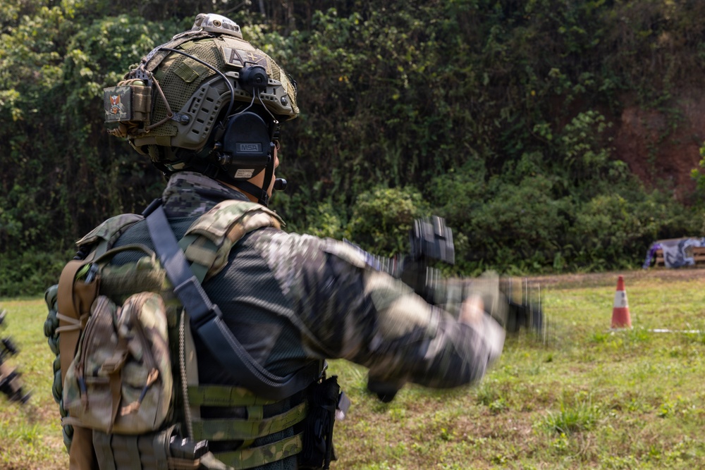 A heavily equipped military or special operations soldier is photographed from behind in an outdoor field environment. The soldier is wearing full tactical gear including a helmet with communications equipment, body armor, and a backpack. Their arm is in motion (motion blur visible), suggesting they are signaling or gesturing toward a training area.