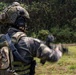 A heavily equipped military or special operations soldier is photographed from behind in an outdoor field environment. The soldier is wearing full tactical gear including a helmet with communications equipment, body armor, and a backpack. Their arm is in motion (motion blur visible), suggesting they are signaling or gesturing toward a training area.
