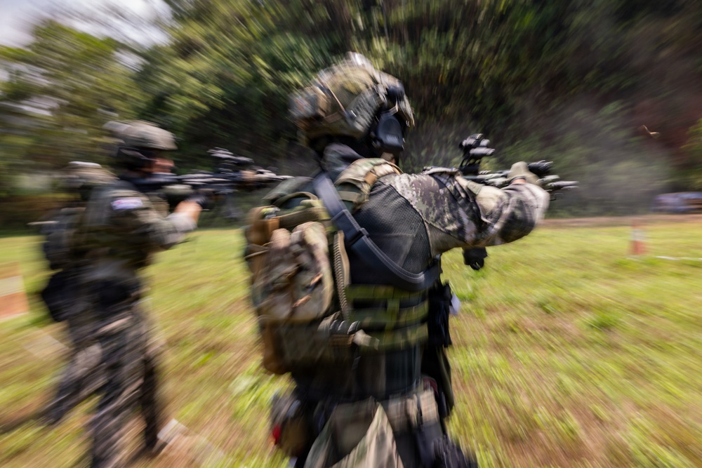 The image shows heavily armed and equipped tactical/military personnel in full combat gear moving through an outdoor area with trees and grass. The photo uses a zoom burst or radial blur technique, creating a dynamic motion effect centered on the foreground soldier. At least two armed individuals are visible, both carrying weapons in a ready/aimed position.