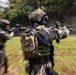 The image shows heavily armed and equipped tactical/military personnel in full combat gear moving through an outdoor area with trees and grass. The photo uses a zoom burst or radial blur technique, creating a dynamic motion effect centered on the foreground soldier. At least two armed individuals are visible, both carrying weapons in a ready/aimed position.