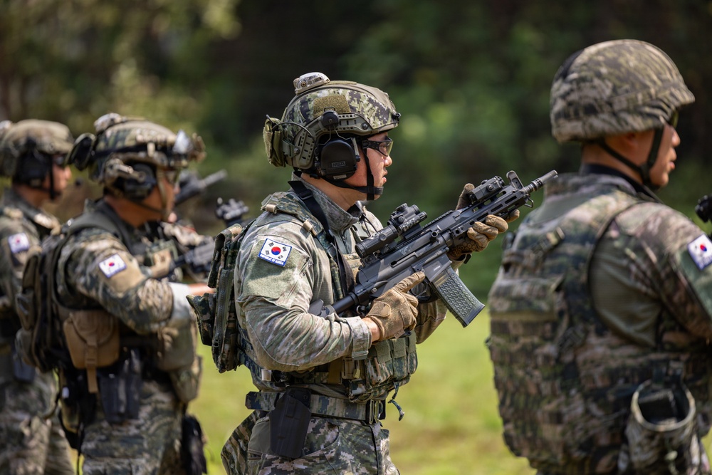 The image shows multiple soldiers in South Korean military uniforms conducting what appears to be a field training exercise or demonstration. The soldiers are equipped with full tactical gear including helmets, body armor, communication headsets, and assault rifles. South Korean national flag patches are clearly visible on their uniforms.