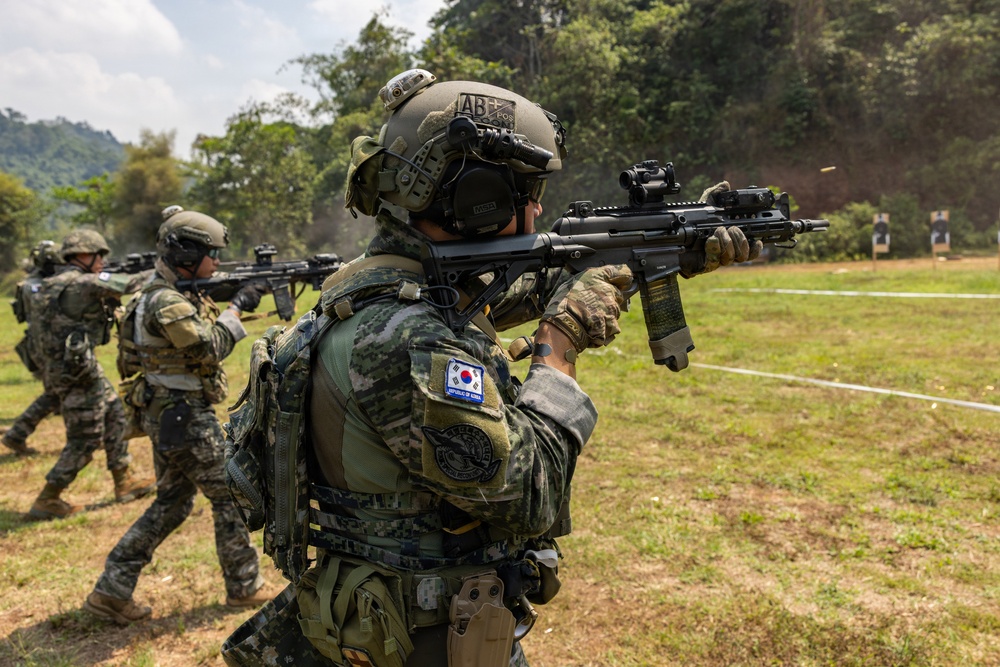 Military personnel are conducting a live-fire shooting exercise on an outdoor range surrounded by vegetation. The foreground soldier wears a 'Republic of Korea' patch and South Korean flag insignia, indicating this is a South Korean military training exercise, possibly a joint operation. Multiple soldiers are actively engaging targets visible on the right side of the frame.