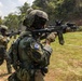 Military personnel are conducting a live-fire shooting exercise on an outdoor range surrounded by vegetation. The foreground soldier wears a 'Republic of Korea' patch and South Korean flag insignia, indicating this is a South Korean military training exercise, possibly a joint operation. Multiple soldiers are actively engaging targets visible on the right side of the frame.