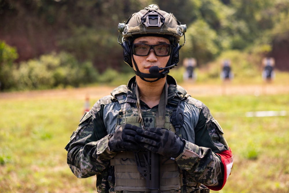 A soldier in full tactical gear stands at what appears to be a military training range or exercise area. The individual is equipped with modern combat equipment including a tactical helmet, communications headset, body armor, and protective eyewear. Shooting targets and other personnel are visible in the blurred background.