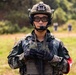 A soldier in full tactical gear stands at what appears to be a military training range or exercise area. The individual is equipped with modern combat equipment including a tactical helmet, communications headset, body armor, and protective eyewear. Shooting targets and other personnel are visible in the blurred background.