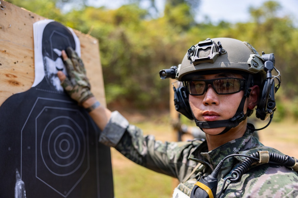A military or law enforcement personnel in full tactical gear is examining a shooting range target. The individual is wearing camouflage uniform, tactical helmet with night vision mount, ballistic eyewear, and communications headset. The setting appears to be an outdoor shooting range with vegetation visible in the background.