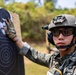 A military or law enforcement personnel in full tactical gear is examining a shooting range target. The individual is wearing camouflage uniform, tactical helmet with night vision mount, ballistic eyewear, and communications headset. The setting appears to be an outdoor shooting range with vegetation visible in the background.