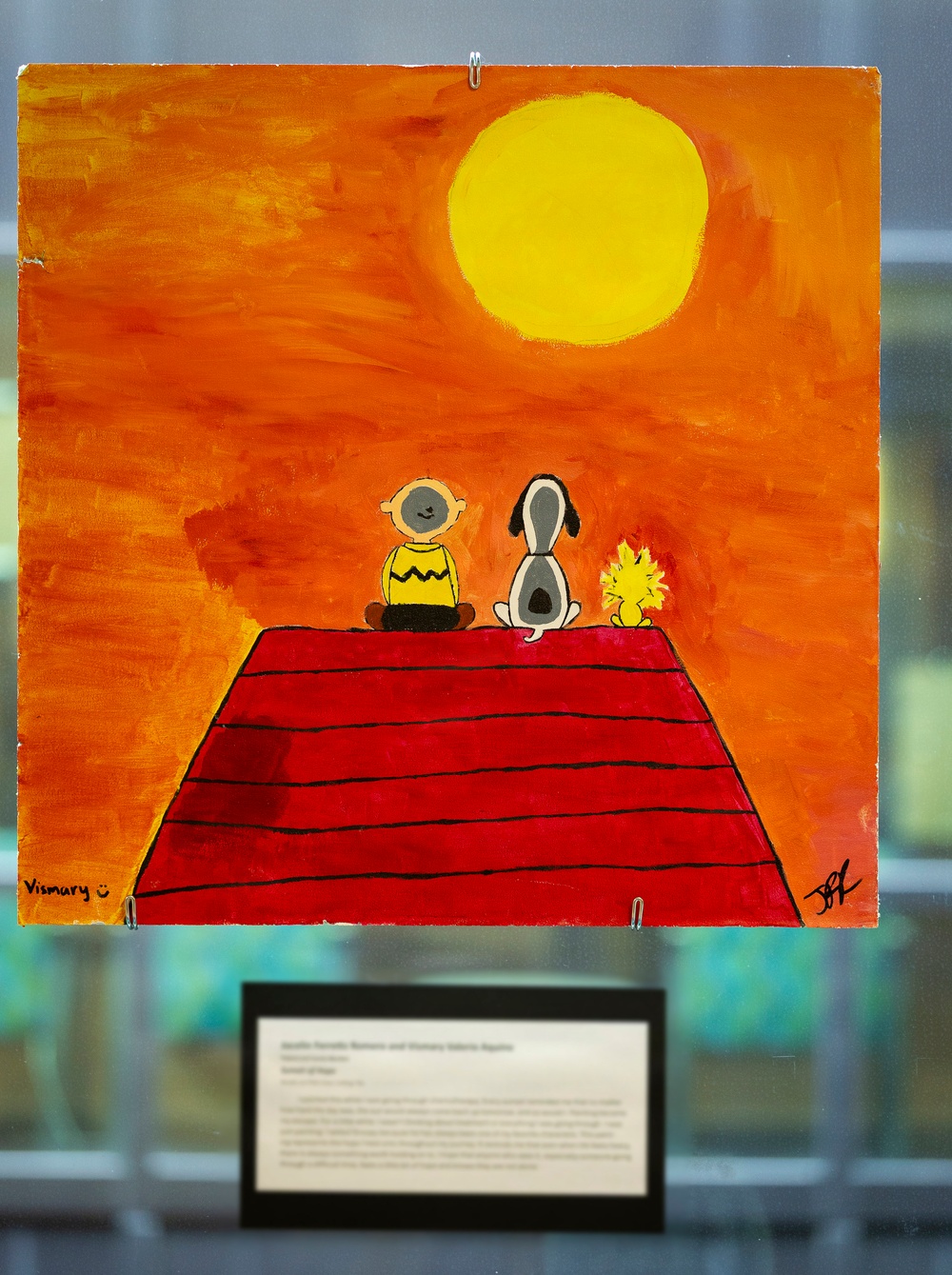 A painted artwork displayed in what appears to be a gallery or exhibition setting, depicting Peanuts characters Charlie Brown, Snoopy, and Woodstock sitting on top of Snoopy's red doghouse against a vivid orange sunset sky with a large yellow sun. The painting is signed 'Vismary' on the lower left and has an additional signature on the lower right. A label placard is visible below the artwork.