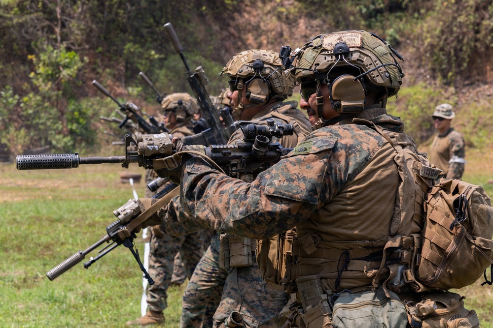A line of military personnel in full combat gear are engaged in a live-fire or training exercise on an outdoor range. The soldiers are wearing MARPAT (Marine Pattern) digital camouflage uniforms, tactical helmets, and hearing protection, and are aiming rifles downrange. A supervising figure in lighter gear is visible in the background to the right.