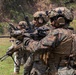 A line of military personnel in full combat gear are engaged in a live-fire or training exercise on an outdoor range. The soldiers are wearing MARPAT (Marine Pattern) digital camouflage uniforms, tactical helmets, and hearing protection, and are aiming rifles downrange. A supervising figure in lighter gear is visible in the background to the right.
