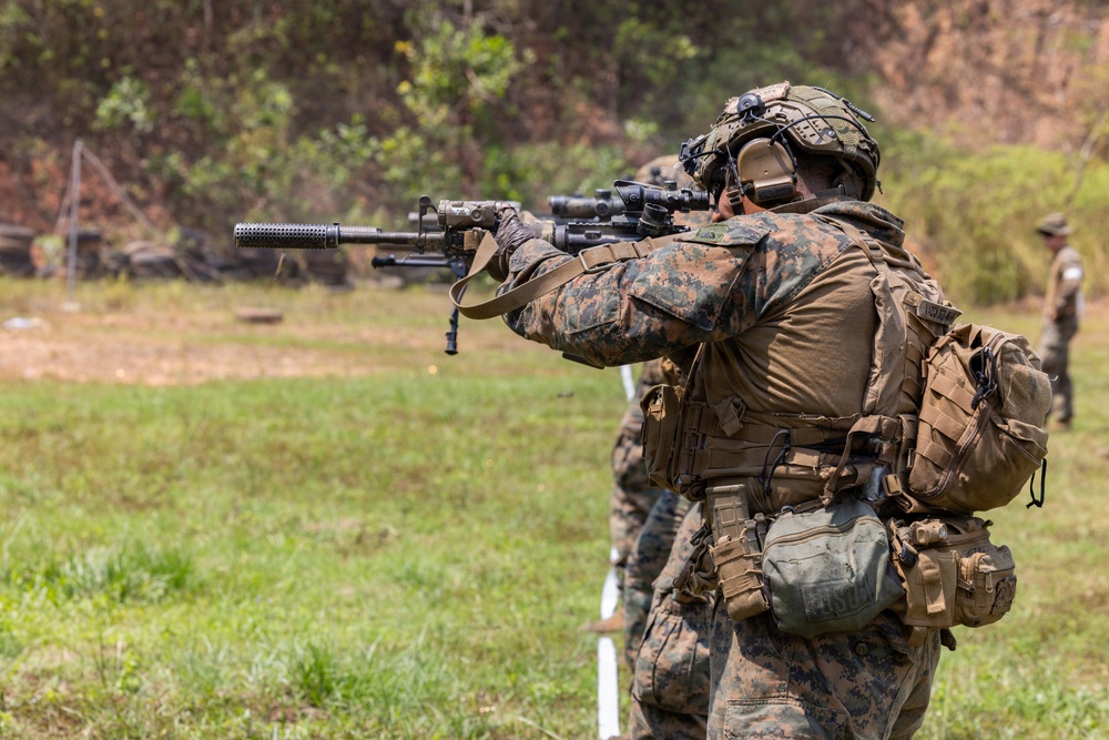 A heavily equipped military service member in MARPAT (Marine digital camouflage) uniform is aiming a rifle fitted with a suppressor and optic scope at a shooting range. The soldier is wearing full tactical gear including a helmet with mounted devices, ear protection, and a loaded plate carrier with multiple pouches. At least one additional service member and a range safety observer are visible in the background.