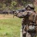 A heavily equipped military service member in MARPAT (Marine digital camouflage) uniform is aiming a rifle fitted with a suppressor and optic scope at a shooting range. The soldier is wearing full tactical gear including a helmet with mounted devices, ear protection, and a loaded plate carrier with multiple pouches. At least one additional service member and a range safety observer are visible in the background.
