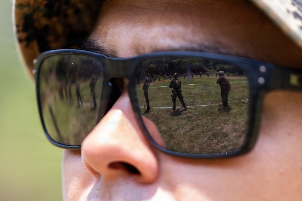 A close-up photograph of a person wearing military camouflage headgear and dark-tinted sunglasses. Reflected in the lenses are multiple armed military personnel standing on an open grassy field, suggesting a training exercise or range activity. The creative composition uses the sunglasses as a mirror to reveal the scene the subject is observing.