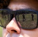 A close-up photograph of a person wearing military camouflage headgear and dark-tinted sunglasses. Reflected in the lenses are multiple armed military personnel standing on an open grassy field, suggesting a training exercise or range activity. The creative composition uses the sunglasses as a mirror to reveal the scene the subject is observing.