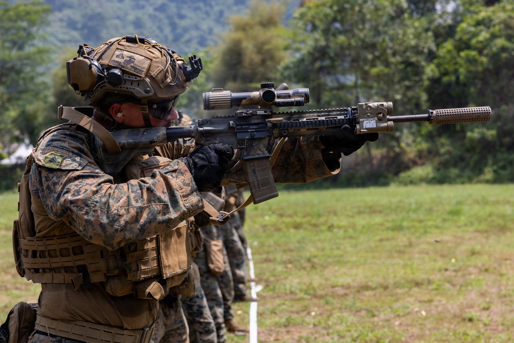 A U.S. Marine in full combat gear is kneeling and aiming a rifle equipped with a scope and suppressor at a shooting range. Additional Marines are visible in the background in a similar firing line formation. The setting appears to be an outdoor training exercise in a tropical or subtropical environment.