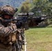 A U.S. Marine in full combat gear is kneeling and aiming a rifle equipped with a scope and suppressor at a shooting range. Additional Marines are visible in the background in a similar firing line formation. The setting appears to be an outdoor training exercise in a tropical or subtropical environment.
