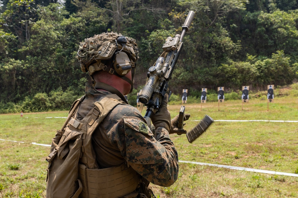 A military service member in full combat gear is performing a magazine reload drill at an outdoor shooting range. The soldier is wearing MARPAT (Marine pattern) camouflage uniform with tactical helmet, hearing protection, and a combat vest. Multiple silhouette paper targets are visible downrange against a treeline backdrop.