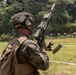 A military service member in full combat gear is performing a magazine reload drill at an outdoor shooting range. The soldier is wearing MARPAT (Marine pattern) camouflage uniform with tactical helmet, hearing protection, and a combat vest. Multiple silhouette paper targets are visible downrange against a treeline backdrop.