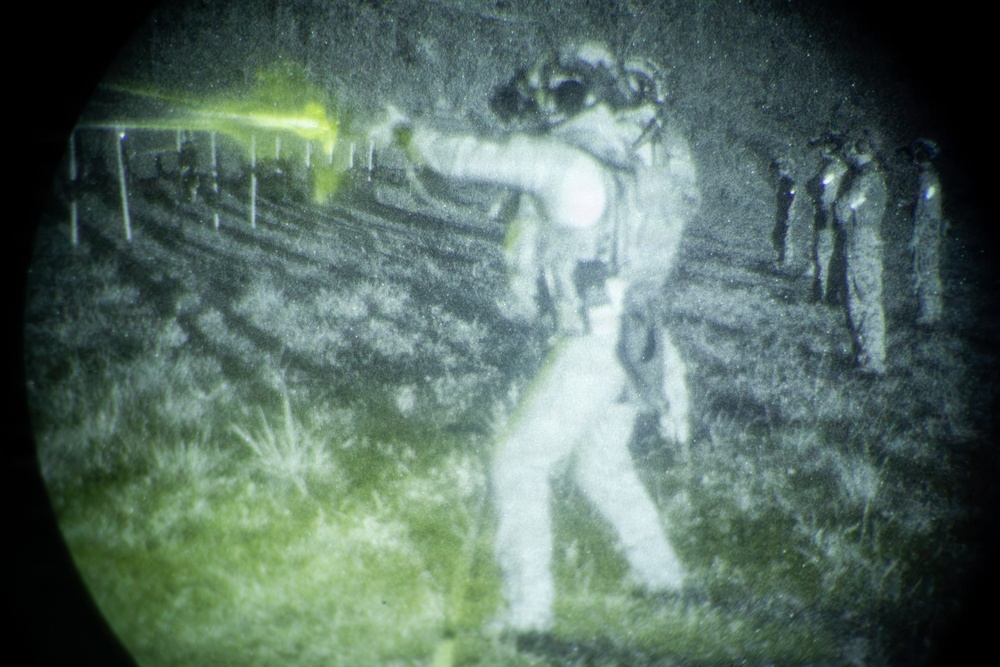 A night-vision captured image showing a military or law enforcement training exercise on a firing range. A central figure in tactical gear is in a shooting stance with a visible laser aiming device emitting a bright green beam toward downrange targets. Additional personnel are visible observing from the right side of the frame.