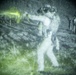 A night-vision captured image showing a military or law enforcement training exercise on a firing range. A central figure in tactical gear is in a shooting stance with a visible laser aiming device emitting a bright green beam toward downrange targets. Additional personnel are visible observing from the right side of the frame.