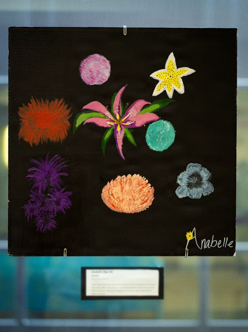 A painting on a black background featuring multiple colorful flowers in various styles, signed 'Anabelle' in the lower right corner with a small yellow flower incorporated into the signature. The artwork is displayed behind glass with a label/placard visible below it. The piece appears to be a student or youth artwork displayed in an exhibition setting.