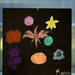 A painting on a black background featuring multiple colorful flowers in various styles, signed 'Anabelle' in the lower right corner with a small yellow flower incorporated into the signature. The artwork is displayed behind glass with a label/placard visible below it. The piece appears to be a student or youth artwork displayed in an exhibition setting.
