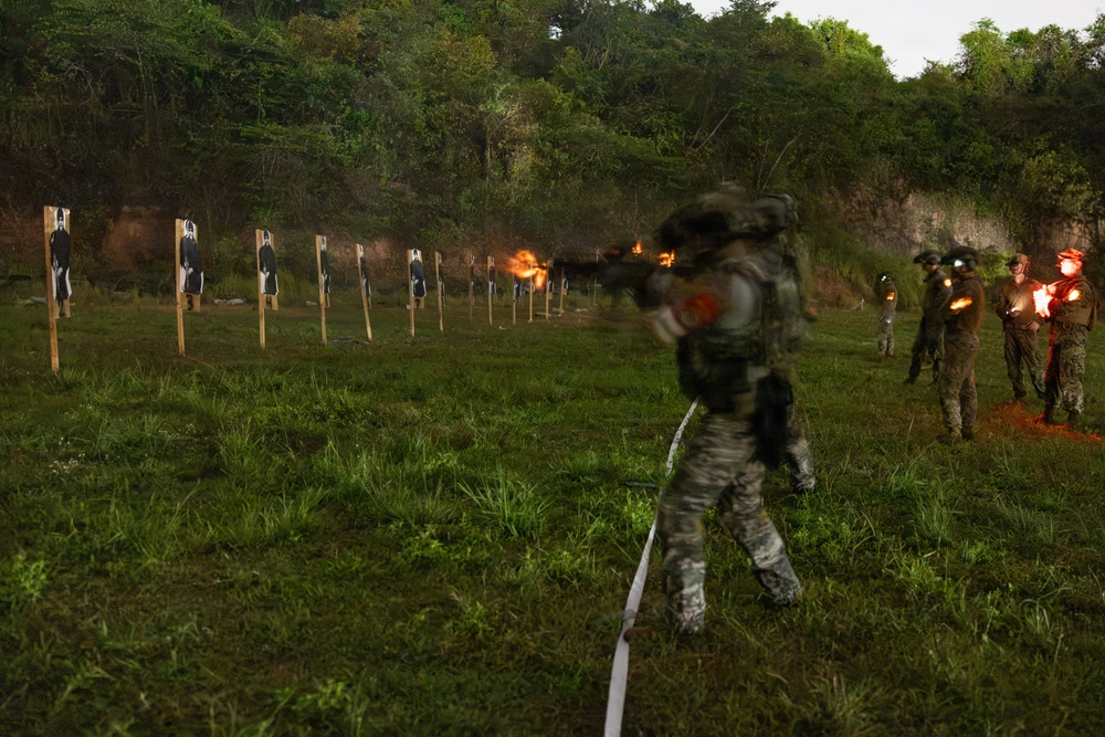 A military or law enforcement training exercise is taking place on an outdoor shooting range at dusk or low-light conditions. A shooter in camouflage gear is moving along a marked lane while firing at a row of paper silhouette targets, with visible muzzle flash captured by the camera. Several other personnel in tactical gear are observing or waiting their turn on the right side of the frame.